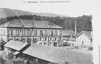 Grand bâtiment blanc à trois étages et toit à quatre pans et petit baraquement en bois ou à colombage surmonté d'une cheminée en brique rouge.