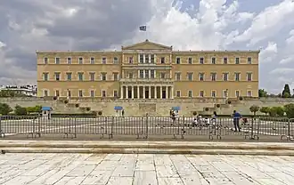 Façade d'un immense bâtiment de pierres jaunes, derrière une vaste place avec des grilles.