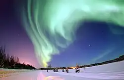 Devant un ciel turquoise, une grande traînée verte est visible. Au sol, on observe de la neige et de sapins.