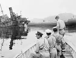 Vue en noir et blanc de l'avant d'une barque avec 4 soldats. En arrière-plan un navire naufragé.