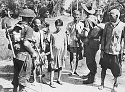 Photo noir et blanc de trois hommes armés en uniforme avec des civils : trois hommes âgés et un adolescent.