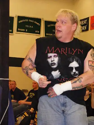 Un homme blond aux bras tatoué portant un t-shirt noir et un jean.