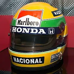 Photographie couleur du casque de Senna, majoritairement jaune, avec une bande verte et des sponsors, au musée Honda.