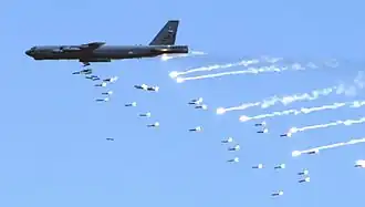 Passage à basse altitude d'un B-52 de couleur très sombre. Sa soute à bombes est ouverte et une nuée de bombes est en train de sortir par cette ouverture.