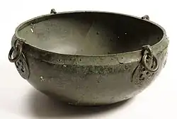 Photo d'un bol en métal gris corrodé. Deux disques en métal ouvragés en décorent la face visible, et des anneaux de métal sont passés dans des crochets fixés à ces disques