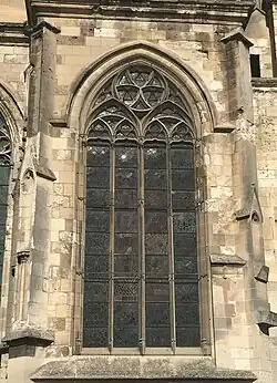 Dans un mur de pierres, encadrée par deux niches, une grande fenêtre composé de quatre lancettes, surmontées par des formes géométriques.
