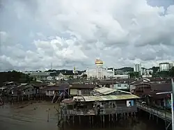 Photographie d'un quartier pauvre de la capitale, Bandar Seri Begawan. Au premier plan se trouvent des maisons sur pilotis délabrées. En arrière-plan se trouve la mosquée Omar Ali Saifuddin et des immeubles modernes en béton.
