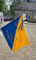 Photo montrant au premier blanc un drapeau de couleur bleu et jaune, à l'arrière des spectateurs sur un trottoir