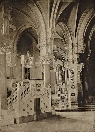 Photographie en noir et blanc montrant l'intérieur d'une église, avec des arcades et une chaire en marbre ainsi qu'un autel à l'arrière-plan.