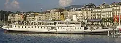 Photographie en couleurs d'un bateau à vapeur blanc devant des immeubles.