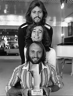 3 hommes souriants aux cheveux longs de face, le menton de l’un posé sur le front de l’autre, 2 d’entre eux portent la barbe