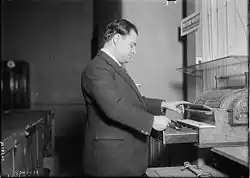 Photographie en noir et blanc d'une personne debout, en costume de couleur sombre, trois quart de profil, regardant et actionnant une caisse enregistreuse.