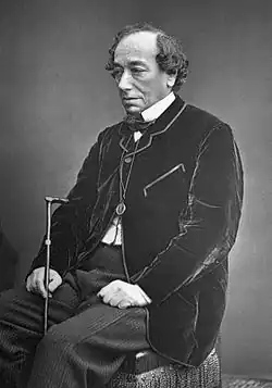 Portrait d'un homme âgé à l'air fatigué assis sur une chaise. Il arbore un petit bouc et a les cheveux bouclés rabattus sur le côté. Il porte une veste noire en velours, un monocle en pendentif et tient une canne.