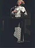 Photo d'une dame âgée dans une longue robe derrière un présentoir avec microphone lors d'une cérémonie.