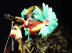 Image d'une femme brune habillée de noir et avec des tours d'oreilles à plumes bleues chantant dans un micro
