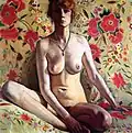 Portrait de face d'une femme nue assise sur un canapé fleuri, visage à moitié dans l'ombre, jambes repliées et bras droit cachant en partie le sexe