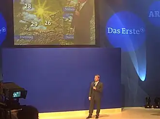 Présentateur météo faisant sa description du temps devant un écran bleu, et en haut l'image vue à la télévision.