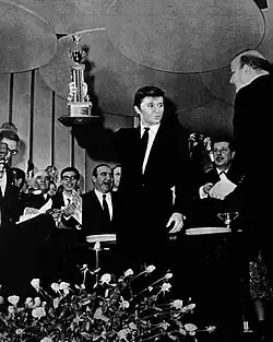 Photographie en noir et blanc d'un jeune homme debout, brandissant de la main droite un trophée : sur la droite, un homme ; au fond une foule de personnages ; devant, un grand bouquet de fleurs.