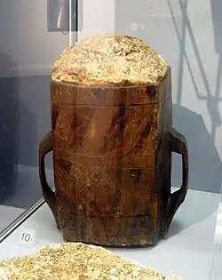Photo d'un récipient en bois foncé, de section quadrangulaire arrondie, à deux poignées, rempli d'une substance jaunâtre.