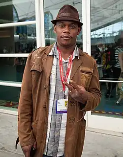 Un homme de face avec un chapeau et une veste en cuir. Il regarde la caméra et fait un V avec deux de ses doigts.