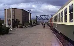 Photographie d'un train arrêté le long d'une plateforme ferroviaire à la fin de laquelle se trouve un pont en arc métallique. Un bâtiment en béton et un panneau portant les insignes de la RDA sont visibles à côté de la plateforme. Plusieurs personnes marchent sur la plateforme et les portes du train sont ouvertes.