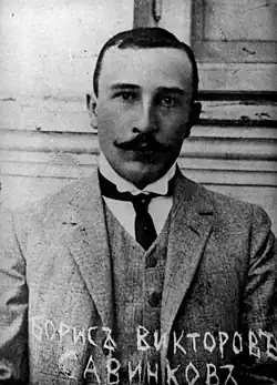 Photo noir et blanc d'un jeune homme, de face et en buste, avec costume trois pièces clair, faux col et cravate, visage sérieux, moustache en croc et front dégarni