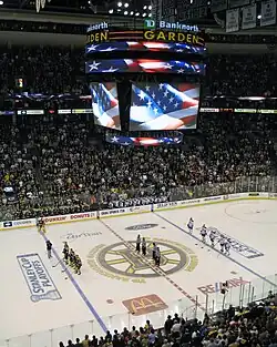 Photo montrant les joueurs des Bruins, les arbitres et les joueurs des Canadiens alignés lors des hymnes sur la patinoire marquée du logo des Bruins.