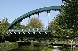  Photo couleurs. Le viaduc de Pannes est vu de côté, sur sa moitié gauche, un arbre au feuillage verdoyant occupe la partie droite de la photo. Le pont est constitué d’un large arc métallique de couleur vert émeraude, sur lequel sont arrimées des barres de fer, appelées suspentes, disposées de manière rayonnante, et qui soutiennent le tablier du pont. Le pont enjambe le canal. Le chemin de halage est à droite de la photo.