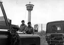 Deux soldats britanniques portant des fusils se tiennent derrière deux jeeps dont l'une porte une plaque « British Frontier Service ». Derrière eux se trouve une haute clôture grillagée et une grande tour de surveillance avec un sommet octogonal.
