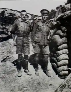 Deux hommes en uniforme militaire se tiennent côte à côte. L'homme de gauche est légèrement plus petit et plus trapu, et l'homme de droite a le bras appuyé sur une pile de sacs de sable.