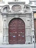 Baie d'imposte ovale à grillage rayonnant coiffée par un larmier droit et cantonnée par deux volutes et de lourdes guirlandes de fruits.