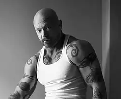 Homme chauve avec une moustache, des tatouages sur le corps et portant un top-tank