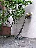Photographie d'une statue en partie dorée, appuyée contre un mur blanc ; à gauche un arbre.