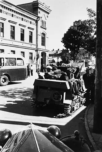 Photo en noir et blanc de 5 ou 6 hommes armés dans un petit véhicule chenillé, sur une avenue urbaine.