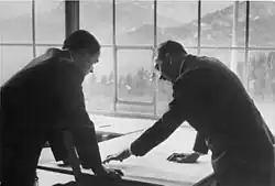 Photo noir et blanc. Adolf Hitler (à droite) et Albert Speer (à gauche), tous deux de profil, en costume sombre, discutent, la tête penchée au-dessus d'une carte étalée sur une plan. En arrière-plan, on voit, à travers une baie vitrée, un paysage de montagne.