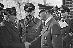 Photo noir et blanc prise le 24 octobre 1940. Au premier plan, Philippe Pétain, chef de l'État français, et Adolf Hitler (tous les deux debout et de profil) échangent une poignée de main. Entre les deux hommes, au second plan, se tient l'interprète officiel de Hitler : Paul-Otto Schmidt. On distingue aussi, au second plan, à droite, le ministre allemand des Affaires étrangères : Joachim von Ribbentrop.