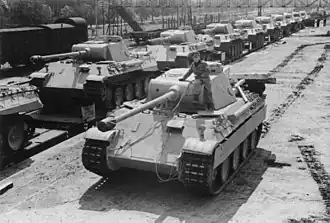 Photographie de 1943 en noir et blanc montrant un char Panther D avançant le long d’un quai de gare, afin d’embarquer sur un train sur lequel sont déjà chargés des dizaines d’autres Panther.