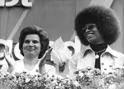 Photographie en noir et blanc de deux femmes derrière des fleurs, l'une blanche avec une coupe de femme traditionnelle, l'autre à sa droite noire avec une coupe afro