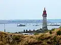 Phare blanc et rouge au sommet ; au premier plan, herbe jaunie ; en arrière plan, des rochers et la mer