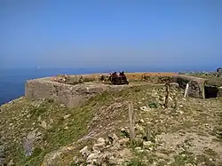 Photographie en couleur montrant un cercle en béton armé (au milieu d'une végétation éparse et très caillouteuse), et au milieu de ce rond les restes en ferraille d'un canon. Le canon est sur une position surélevée et ciel bleu est dégagé.
