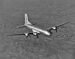 Vue en contre-plongée d'un gros avion quadrimoteur survolant les plaine californiennes ; on peut distinguer au sol une exploitation de puits de pétrole avec une raffinerie à proximité, des champs et une zone résidentielle.