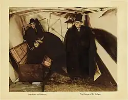 Une lobby card avec une photographie au centre, et de petites légendes en dessous. L'image représente un homme vêtu d'un manteau sombre, d'une cape et d'un chapeau haut de forme, debout à droite, tandis que deux hommes enquêtent sur un autre homme assis dans une boîte ouverte.