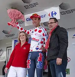 De trois-quarts, trois personnes sourient. Un coureur portant un maillot blanc à pois rouges soulève un bouquet rose.