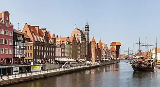 Photographie couleur présentant le panorama d'un quai que longent des bâtiments colorés et une grande grue de bois, ainsi qu'un vaisseau.