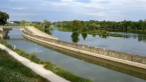  Photo représentant le bief de Saint-Jean-de-Braye en premier plan avec la Loire en second plan. Cette photo met en évidence la particularité de ce bief, à savoir son aspect latéral à la Loire. Il n’est séparé du fleuve que par une sorte de digue constituée de deux murs de soutènement. Au fond de la photo, on voit un pont en treillis métallique permettant aux piétons de franchir le canal. Au premier plan figure le chemin de promenade, ancien chemin de halage