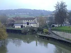 Une rivière coule lentement à partir du milieu à gauche vers le bas à droite, avec l'entrée d'un canal vers le coin supérieur droit. Le canal passe immédiatement sous un petit pont, et à 100 mètres plus loin, sous un autre pont. Une colline avec des maisons forment l'arrière-plan.