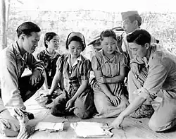 Groupe de femmes assises en tailleur devant des documents. Deux hommes leur parlent.