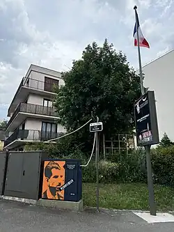 Photographie d'un angle de rue. Au centre, on peut apercevoir une armoir électrique sur laquelle le visage de Missak Manouchian a été peint.