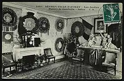 Carte postale en noir et blanc oblitérée d'un timbre vert montrant l'intérieur d'un salon comprenant des tableaux et des portraits photographiques accrochés au mur, ainsi que des objets d'art disposés sur des meubles et une petite cheminée.