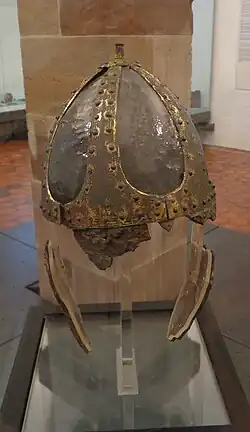 Photographie d'un casque pointu en métal, plutôt abîmé et présenté dans une vitrine d'un musée.